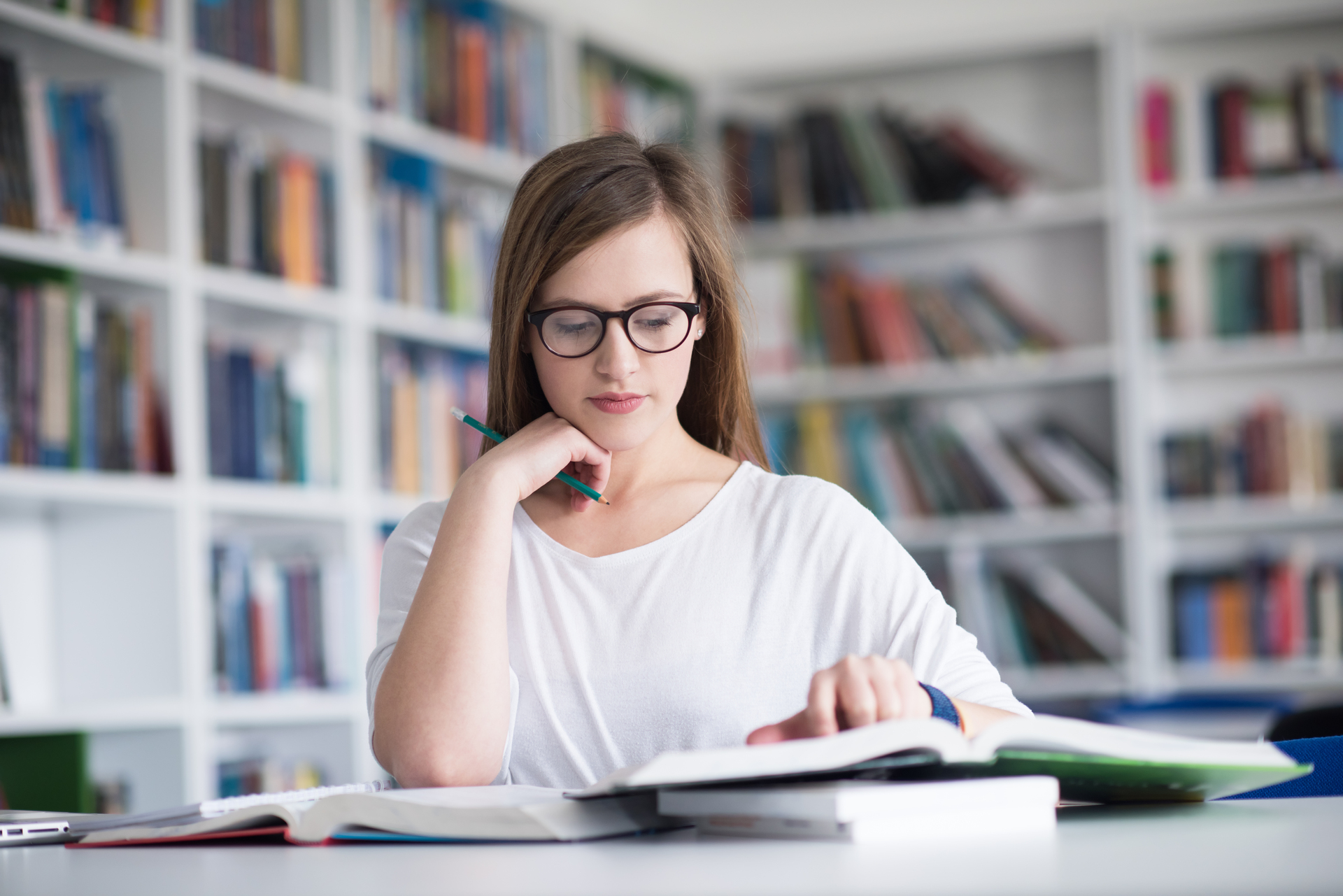 A female teenage student with brunette hair and glasses studying at a desk in a library with an open book in front of her.