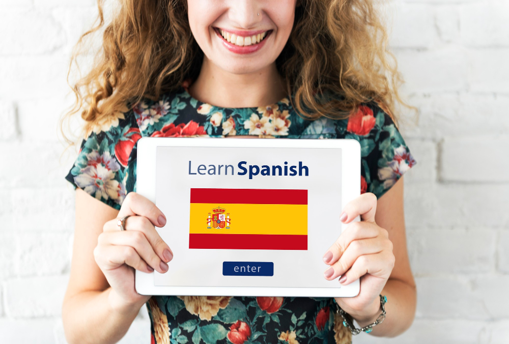 A smiling girl holds a sign featuring the Spanish flag and the words “Learn Spanish”.