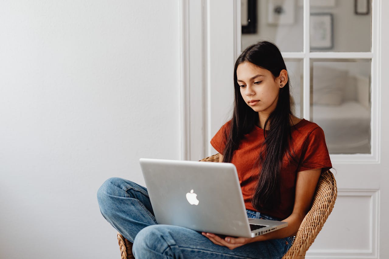 Teenage girl with long brunette hair in red t-shirt using Apple laptop, sitting down and looking curious.