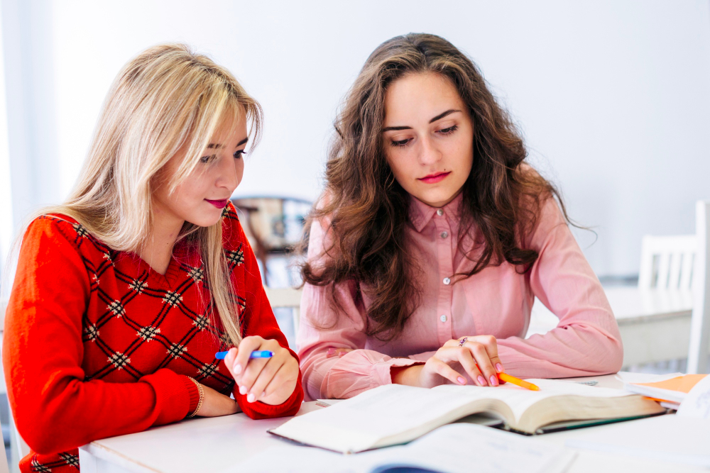 Two teenage girls studying together at a desk with an open book and pens.