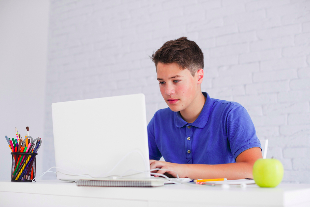Teenage student in blue polo studying with laptop at desk.